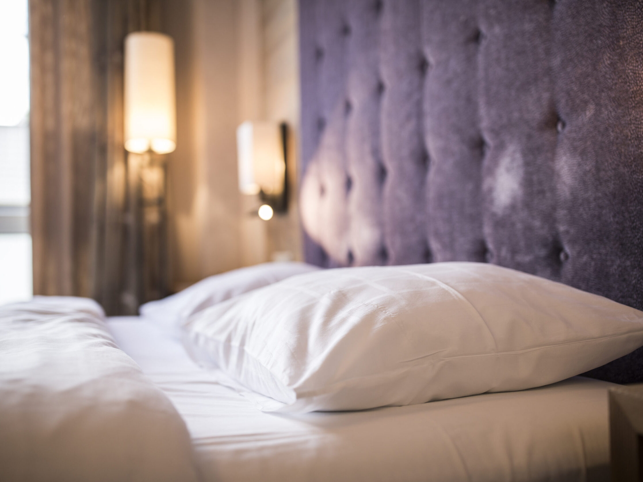 Detailansicht eines komfortablen Hotelbetts mit lila Stoffkopfteil und gemütlicher Beleuchtung.