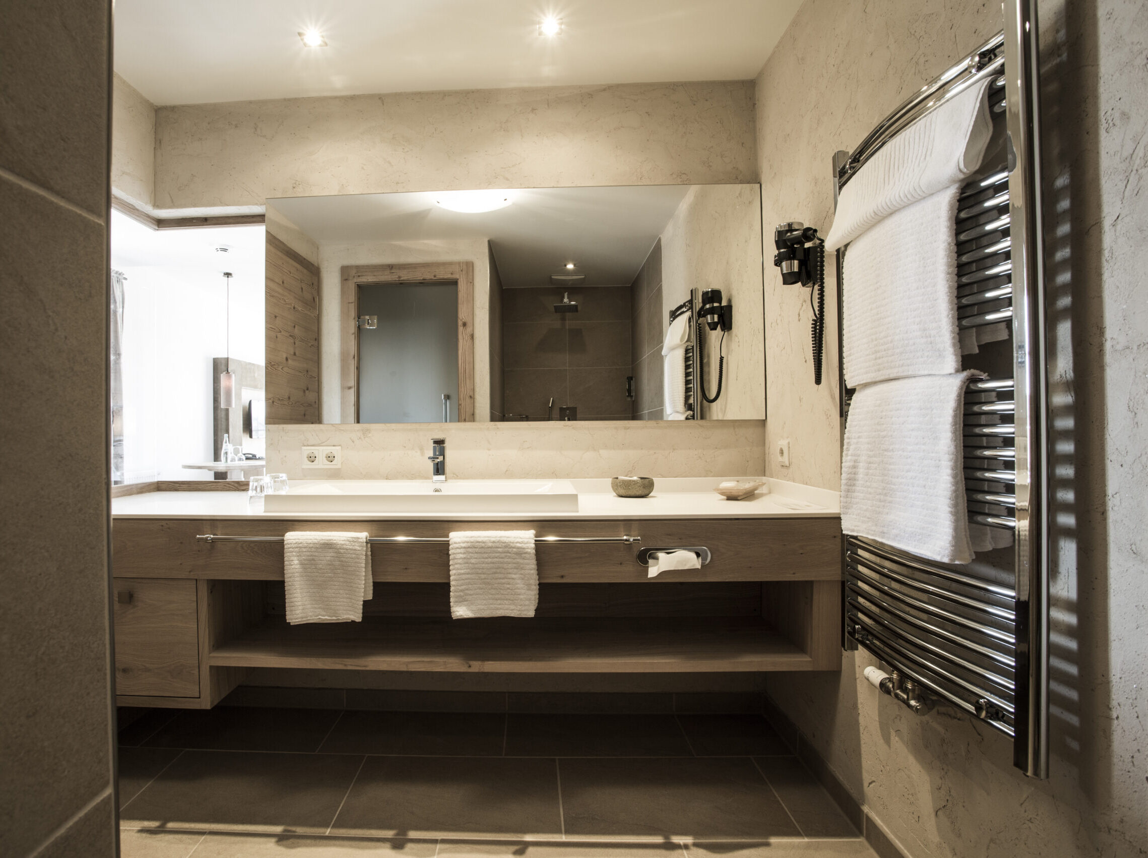Junior Suite Superior Plus mit zwei Balkonen im Meiser SPA Resort. Mit Blick ins Badezimmer, XXL Regendusche, großer Spiegel und elegantem Holzdesign